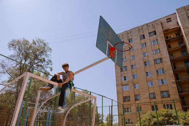 Teenage boy sit on top of a soccer net with a basketball hoop and apartment building behind him. He looks confident and aloof.